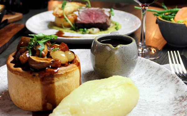A photo of a steak pie and mashed potato on a plate with a small jug of gravy. A glass of red wine, another plate of food can be seen in the background and a bowl of vegetables can be seen on the back right.