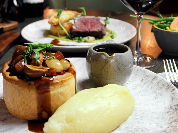 A photo of a steak pie and mashed potato on a plate with a small jug of gravy. A glass of red wine, another plate of food can be seen in the background and a bowl of vegetables can be seen on the back right.