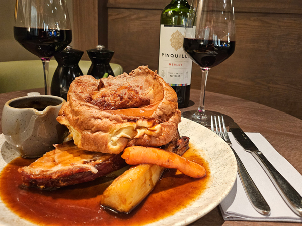 A photo of a roast diner on a plate, including a large Yorkshire pudding, a roasted carrot and parsnip, and a small rug of gravy. A knife and fork lay on a folded napkin to the right hand side and two glasses of red wine can be seen in the background along with the bottle and salt and pepper mills.