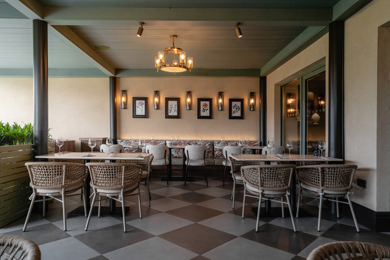 A view of the indoor terrace area at Cook House Bar & Kitchen, Leeds. There is banquette seating along the rear wall with 3 tables and 5 chairs. To the front of the image there two marble tables with  seating for 4.  The floor is tiled in a grey and white chequerboard style, the ceiling is wood-panelled and supported by metal pillars.